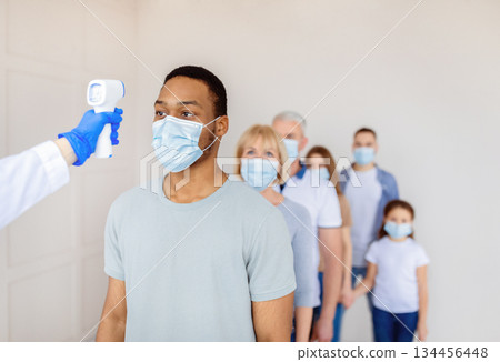 Coronavirus immunization medical campaign concept. Black man in face mask standing in line at clinic, doctor measuring his temperature with infrared thermometer before covid-19 vaccination, copy space 134456448