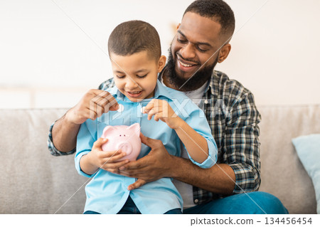Personal Savings And Budget Planning. African American Daddy And His Son Boy Putting Coin In Piggybank Raising Money Sitting On Couch At Home. Child And Financial Literacy 134456454