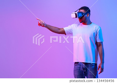 Young Black Man In VR Headset Touching Air While Playing Video Game, Millennila African American Guy Enjoying Virtual Reality Experience, Standing Under Neon Lighting Over Purple Background, Panorama 134456456