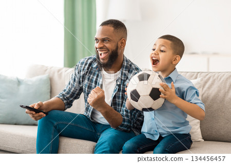 Happy African American Father And Son Watching Sports On TV Holding Soccer Ball And Remote Control Sitting On Sofa At Home. Family Watches Television. Sport TV Programming Concept 134456457