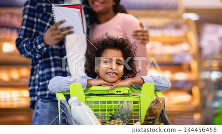 Adorable black girl with full shopping cart and her parents checking grocery list at modern supermarket. Millennial African American family with cute child doing weekly purchases at mall Adorable black girl with full shopping cart and her parents checking grocery list at modern supermarket. Millennial African American family with cute child doing weekly purchases at mall 134456458