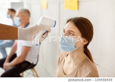 Young woman in face mask waiting for covid-19 immunization at hospital, doctor checking her temperature with infrared thermometer. Vaccination against coronavirus concept Young woman in face mask waiting for covid-19 immunization at hospital, doctor checking her temperature with infrared thermometer. Vaccination against coronavirus concept 134456459