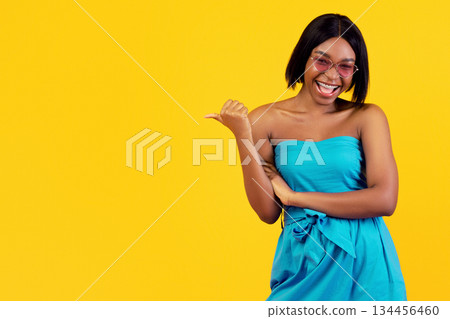 Happy black woman in sexy dress and sunglasses pointing aside or showing hitchhiking gesture on orange studio background. Cheerful lady recommending summertime travel, gesturing thumb up Happy black woman in sexy dress and sunglasses pointing aside or showing hitchhiking gesture on orange studio background. Cheerful lady recommending summertime travel, gesturing thumb up 134456460