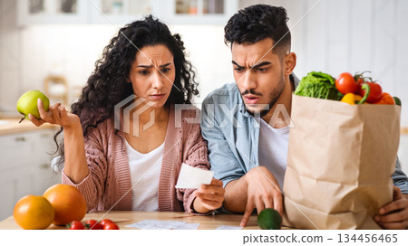 Shocking Prices. Confused Middle Eastern Spouses Checking Bills After Grocery Shopping, Young Arab Couple Sitting At Table In Kitchen, Upset Man And Woman Reading Supermarket Invoice, Closeup 134456465
