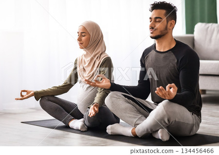 Expecting Muslim Spouses Practicing Yoga Together At Home, Sitting In Lotus Pose With Closed Eyes On Mat In Living Room, Calm Islamic Couple Enjoying Healthy Lifestyle During Pregnancy, Free Space 134456466