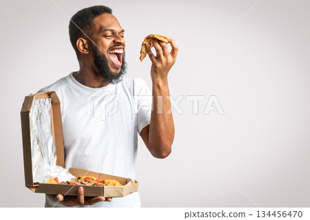 Hungry Black Guy Eating Pizza Slice Standing Over White Studio Background. Fast Food Eater Enjoying Pizza. Junk Food Overeating Habit And Unhealthy Nutrition, Cheat Meal Concept 134456470