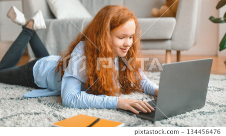 Browsing Internet. Portrait of red-haired girl lying on the floor carpet in living room, using laptop computer, watching video or movie, chatting with friends, surfing web and smiling Browsing Internet. Portrait of red-haired girl lying on the floor carpet in living room, using laptop computer, watching video or movie, chatting with friends, surfing web and smiling 134456476