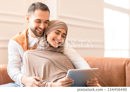 Cheerful Muslim Couple Using Digital Tablet Watching Movie Online Relaxing Sitting And Embracing On Couch At Home On Weekend. Arab Family Browsing Internet Together Indoor 134456490