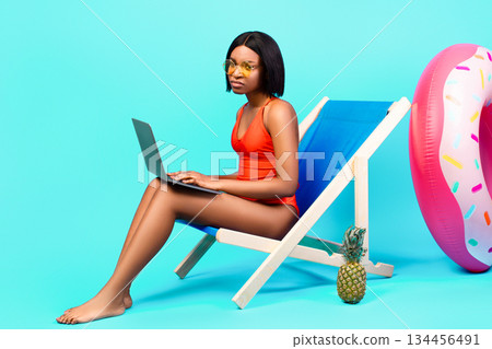 Online work during summer vacation. Unhappy black lady in swimsuit sitting in lounge chair with laptop, having to do urgent job on holidays, participating in business meeting, blue background 134456491