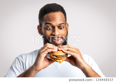 Hungry Black Man Holding Tasty Burger Standing And Licking Lips Posing On White Studio Background. Unhealthy Nutrition And Junk Food Overeating Habit, Cheat Meal Concept Hungry Black Man Holding Tasty Burger Standing And Licking Lips Posing On White Studio Background. Unhealthy Nutrition And Junk Food Overeating Habit, Cheat Meal Concept 134456537