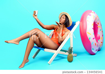 Flirty black woman chatting with her boyfriend on smartphone, blowing air kiss at webcam, making selfie while sitting in lounge chair on beach, blue studio background. Full length portrait 134456541