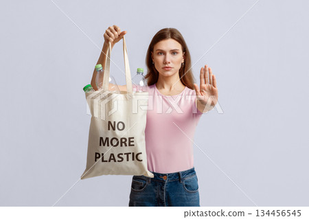 Woman holding eco shopping bag with no more plastic inscription filled with bottles and showing stop gesture with open palm, environmental activist standing over light background in studio 134456545