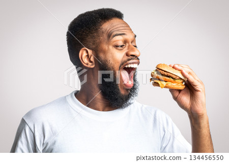 Side View Of Hungry African American Guy Eating Burger Standing In Studio Over White Background. Junk Food And Overeating Habit. Black Man Biting Cheeseburger Unhealthy Nutrition Concept 134456550