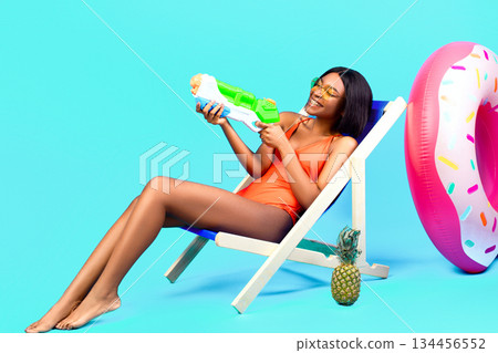 Summertime fun. Attractive black woman sitting in lounge chair with water gun, playing beach game over blue studio background, full length. Happy young lady chilling on tropical resort 134456552