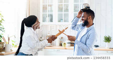 Joyful black couple having fun in kitchen, playfully fighting with kitchenware, aiming each other with utensils tools and laughing, young african spouses having enjoying time at home, copy space Joyful black couple having fun in kitchen, playfully fighting with kitchenware, aiming each other with utensils tools and laughing, young african spouses having enjoying time at home, copy space 134456555