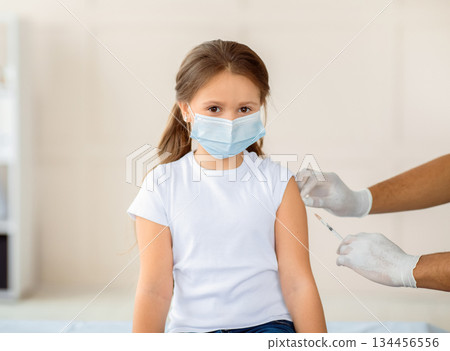 Little girl in face mask being vacciniated against covid-19 at hospital. Cute kid getting coronavirus immunization at health centre. Protection against contagious disease concept Little girl in face mask being vacciniated against covid-19 at hospital. Cute kid getting coronavirus immunization at health centre. Protection against contagious disease concept 134456556