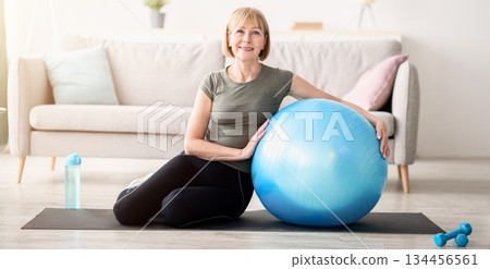 Full length portrait of happy mature woman resting on yoga mat with fitness ball, smiling at camera indoors, free space. Cheerful senior lady taking break from her domestic training 134456561
