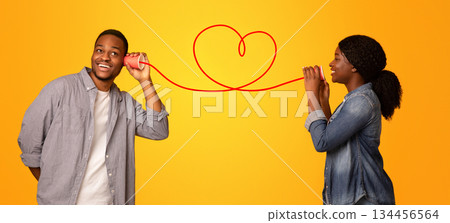 Love Message. Happy black woman whispering romantic words to her boyfriend via tin phone, standing isolated over yellow background, creative collage for Valentine's day with drawn red heart, panorama 134456564