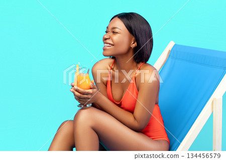 Cheerful young black woman relaxing in lounge chair with delicious fruit cocktail, laughing and enjoying her tropical summer vacation on blue studio background, banner design with copy space 134456579