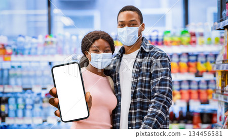 Portrait of black couple in protective masks showing smartphone with empty screen at mall, mockup for design. Young millennial family promoting new groceries list mobile app Portrait of black couple in protective masks showing smartphone with empty screen at mall, mockup for design. Young millennial family promoting new groceries list mobile app 134456581