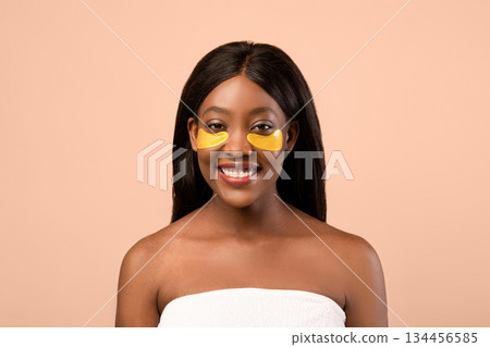 Cheerful attractive young black woman wrapped in towel using golden hydrogel eye patches over beige studio background, copy space. Smiling fair-haired african american lady making eyes care Cheerful attractive young black woman wrapped in towel using golden hydrogel eye patches over beige studio background, copy space. Smiling fair-haired african american lady making eyes care 134456585