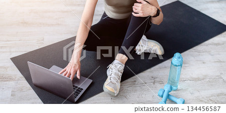 Senior woman watching online sports course on laptop before domestic workout, indoors. Above view of mature lady taking break from exercising, resting on yoga mat with pc. Active lifestyle concept 134456587