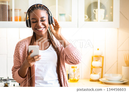 Favorite Leisure. Positive Black Millennial Woman In Headphones Listening Music On Smartphone In Kitchen Interior, Cheerful Young African Female Browsing Modern App With Her Favorite Songs, Copy Space 134456603