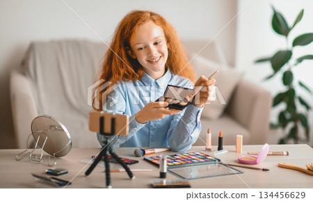 Live Streaming And Blogging Concept. Portrait of smiling teenage girl doing make up, showing her eyeshadow palette, looking at camera, shooting and filming vlog using cellphone on tripod 134456629