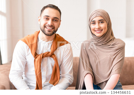 Cheerful Muslim Couple Sitting On Sofa Smiling To Camera In Living Room At Home. Portrait Of Modern Arab Family Of Two, Happy Young Husband And Wife In Hijab Posing Together Indoor Concept 134456631