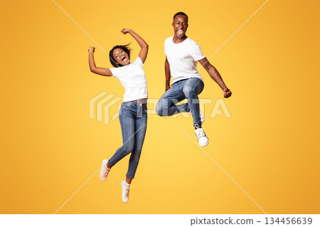 Emotional black millenial couple jumping up on orange studio background, full length shot. Young cheerful african american man and woman celebrating success, having fun together 134456639