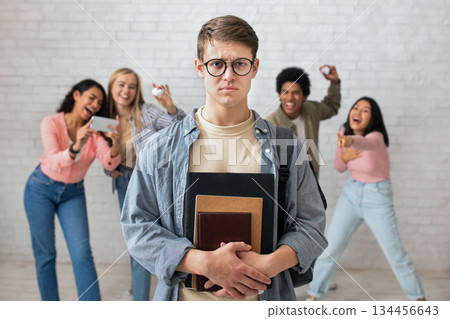 Humiliation and bullying at school. Focus on sad young guy in glasses and notebooks, and multiracial students filming video and throwing papers at guy and laugh at pupil, free space, studio shot 134456643