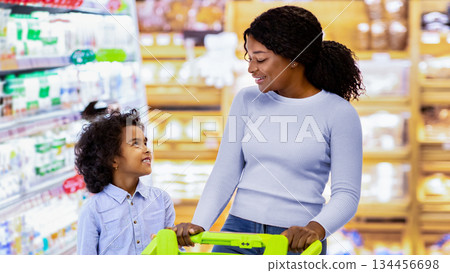 Young black mom and her daughter shopping together for groceries, choosing dairy products at store. Millennial African American family with cart purchasing food at big hypermarket 134456698