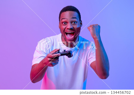 Overjoyed Black Man With Joystick In Hand Celebrating Win In Video Game, African Guy Exclaiming With Excitement And Raising Fist While Standing In Neon Light Over Purple Background, Copy Space Overjoyed Black Man With Joystick In Hand Celebrating Win In Video Game, African Guy Exclaiming With Excitement And Raising Fist While Standing In Neon Light Over Purple Background, Copy Space 134456702