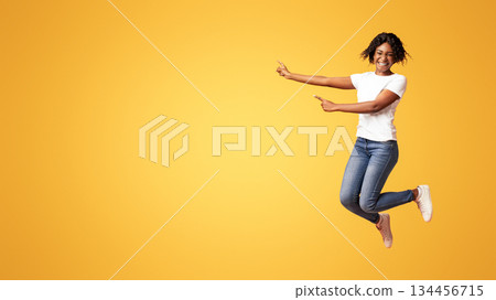 Emotional african american millennial lady smiling and pointing at advertising or announcement. Excited black woman jumping up and showing copy space over orange studio background 134456715