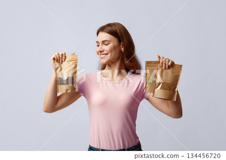 Happy young woman holding brown kraft paper doypack bags with weight groceries, enjoying eco-friendly shopping, smiling millennial lady standing over grey studio background, copy space Happy young woman holding brown kraft paper doypack bags with weight groceries, enjoying eco-friendly shopping, smiling millennial lady standing over grey studio background, copy space 134456720