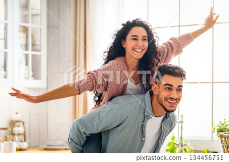 Joyful Middle Eastern Spouses Having Fun Together In Kitchen Interior, Handsome Arab Man Piggybacking His Cheerful Wife, Happy Attractive Lady Enjoying Ride, Spreading Hands Like Flying, Copy Space Joyful Middle Eastern Spouses Having Fun Together In Kitchen Interior, Handsome Arab Man Piggybacking His Cheerful Wife, Happy Attractive Lady Enjoying Ride, Spreading Hands Like Flying, Copy Space 134456751