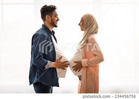 Pregnant islamic lady in hijab comparing belly sizes with her husband, having fun together at home. Joyful middle eastern guy with big fake tummy standing near window in front of her expectant wife 134456758