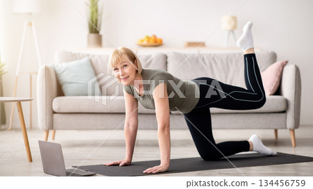 Online home sports concept. Athletic senior woman working out in front of laptop computer in living room. Fit mature lady doing domestic fitness, exercising on yoga mat, strengthening her muscles Online home sports concept. Athletic senior woman working out in front of laptop computer in living room. Fit mature lady doing domestic fitness, exercising on yoga mat, strengthening her muscles 134456759