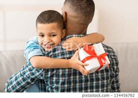 Kid's Birthday. African Little Son Hugging Dad Receiving B-Day Gift Sitting Together On Sofa At Home. Family Holiday, Father's Day Celebration. Gifts And Presents Concept Kid's Birthday. African Little Son Hugging Dad Receiving B-Day Gift Sitting Together On Sofa At Home. Family Holiday, Father's Day Celebration. Gifts And Presents Concept 134456762