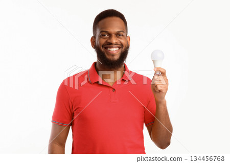 Happy Black Guy Showing Energy-Saving Lightbulb Standing Over White Studio Background, Posing Smiling To Camera. Save Energy, Light Bulb Disposal Service Concept 134456768