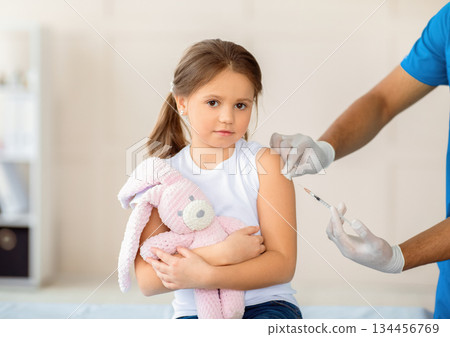 Lovely little girl with toy rabbit receiving covid-19 vaccine injection at clinic. Cute kid getting coronavirus immunization at hospital. Protection against global virus concept 134456769