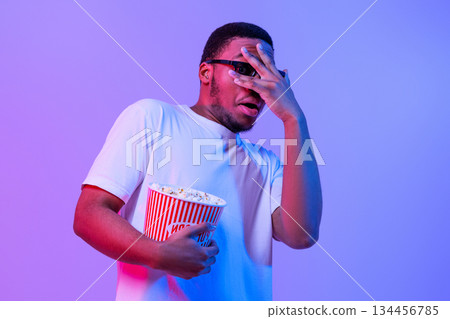 Scary Movie. Thrilled Black Guy In 3D Glasses Watching Horror Film, Millennial African American Man Holding Bucket With Popcorn And Covering Eyes With Afraid, Standing In Neon Purple Light, Free Space 134456785