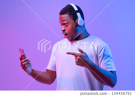 Amazed Black Guy In Wireless Headphones Pointing At Smartphone In His Hands, Excited African American Man Emotionally Reacting To Mobile Offer, Standing In Neon Lighting Over Purple Background 134456791