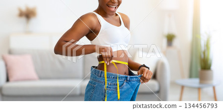 Successful Weight Loss. Joyful Young Black Lady Measuring Waist Wearing Oversized Jeans After Slimming, Smiling To Camera Standing Indoors. Weight-Loss Comparison And Motivation Successful Weight Loss. Joyful Young Black Lady Measuring Waist Wearing Oversized Jeans After Slimming, Smiling To Camera Standing Indoors. Weight-Loss Comparison And Motivation 134456793