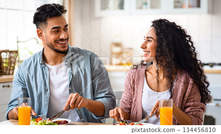 Happy young arab couple eating their breakfast together. Smiling middle eastern spouses sitting at table in kitchen and enjoying tasty healthy meal, spending morning time at home, free space 134456810