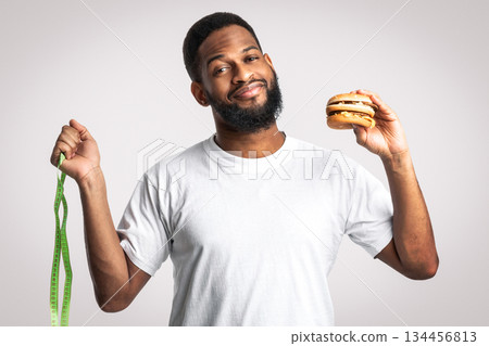 Healthy Vs Unhealthy Lifestyle. African American Man Choosing Between Burger And Skipping Rope Standing Over White Studio Background. Sport Exercise Or Junk Food Choice Concept 134456813