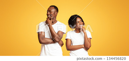 Planning future together. Young african-american couple day dreaming on orange studio background. Pensive black man and woman standing next to each other and thinking, touching their chins 134456820