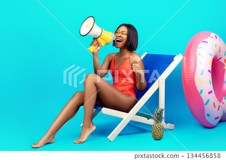 Unbelievable offer. Young black woman in swimwear sitting in lounge chair, having beach vacation, shouting into megaphone, making announcement, advertising your product or service, blue background 134456858