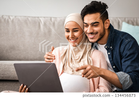 Cheerful Pregnant Muslim Woman In Hijab And Her Husband Using Laptop At Home Together, Middle Eastern Parents Shopping Online With Computer Or Watching Movie, Man Pointing At Screen, Closeup 134456873