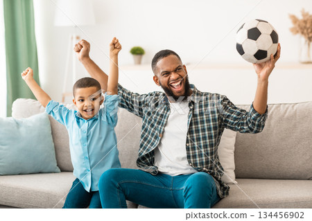 Joyful Black Daddy And Kid Son Watching Soccer Football Game On TV Shaking Fists Cheering Victory Of Favorite Team At Home. Sport Channel Programming Advertisement 134456902